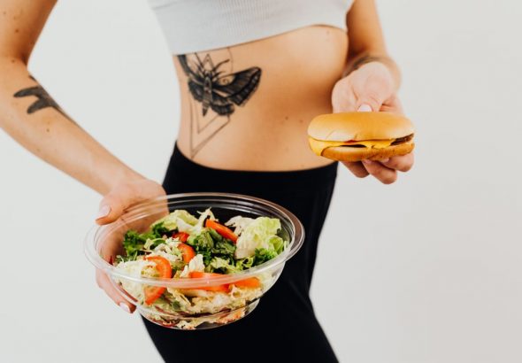 A woman with a tattoo holding a healthy vegetable salad and a cheeseburger, representing food choices.